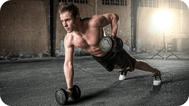 A muscular man performs a plank row with dumbbells in an industrial-style gym setting.